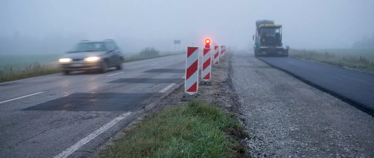 Straßenbaustelle im Morgendunst: Auf der alten, ausgebesserten Fahrbahn fährt ein Auto, direkt daneben entsteht eine neue Fahrbahn aus Schotter und frischem Asphalt.