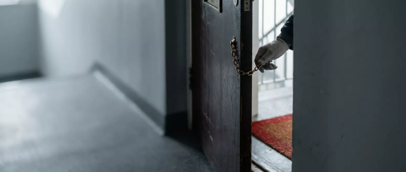 Eine Wohnungstür ist nur einen Spalt breit geöffnet und mit einer Türkette gesichert, eine Hand liegt an der Türkante.
