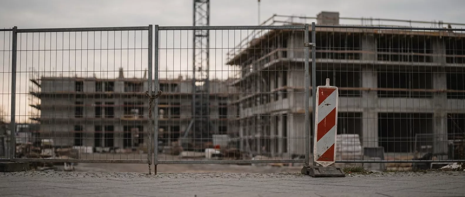 Ein geschlossener Bauzaun vor einem unfertigen Gebäude, dahinter Gerüst und Kran.