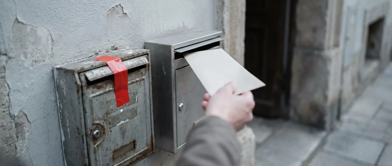 Zwei Briefkästen nebeneinander: Der alte ist mit rotem Klebeband verschlossen, eine Hand wirft einen Brief in den neuen.